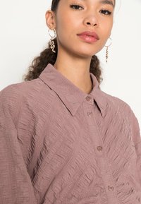 Camisa texturizada rosa com colarinho e fecho de botão, apresentando padrões diagonais elevados. Modelo usa brincos em forma de cobra dourados.