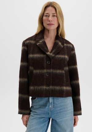 Femme aux longs cheveux blonds porte un manteau en laine à carreaux marron foncé et beige avec des boutons noirs et un jean bleu clair sur un fond uni.