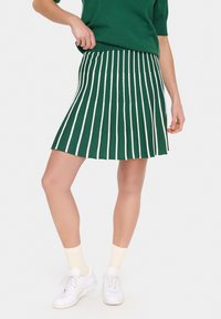 Jupe plissée verte avec des rayures verticales blanches, longueur genou, fabriquée en tissu extensible. Portée avec des baskets blanches et des chaussettes légères.