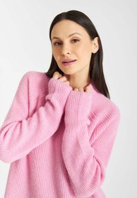 Jasnoróżowy sweter dzianinowy o rypsowym wzorze, z długimi rękawami i okrągłym dekoltem. Materiał wydaje się miękki i lekko puszysty; rękawy zakrywają dłonie.