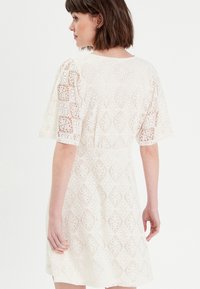 Robe en dentelle blanche à manches courtes, présentant un motif géométrique et une jupe évasée. Le dos met en avant un col arrondi et une taille marquée.