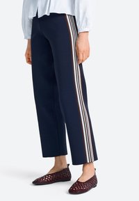 Pantalones anchos azul marino con un diseño lateral de tres rayas blancas y una marrón. Combinados con zapatos planos de tejido en tono burdeos oscuro.