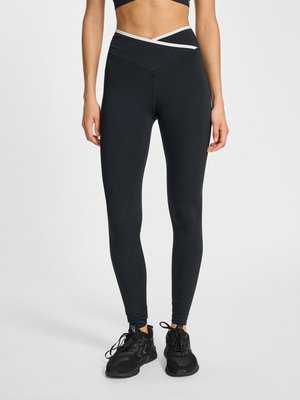 Vrouw met zwarte high-waist legging met wit taillebanddetail en zwarte sportschoenen, staand tegen een effen lichte achtergrond.
