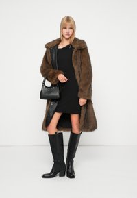 Brūns mākslīgās ādas apmetnis ar ādas akcentiem, melna pieguļoša kleita, melna somiņa ar kniedēm un augstpapēžu melni zābaki līdz ceļiem.