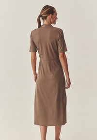 Kurzes braunes Kleid mit kurzen Ärmeln, hohem Ausschnitt und tailliertem Schnitt, gefertigt aus einem glatten, elastischen Stoff, der bis zur Mitte der Wade reicht.