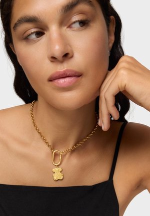 Collar de oro con un charm de oso, presentando un diseño de cadena y un cierre de clip. Textura suave sobre una blusa negra.