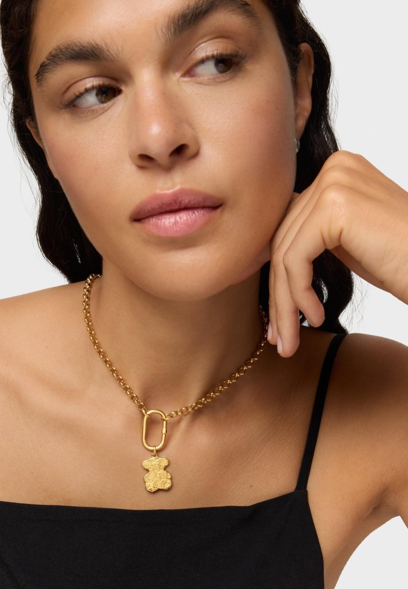 Collar de oro con un charm de oso, presentando un diseño de cadena y un cierre de clip. Textura suave sobre una blusa negra.