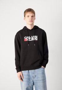 Svart hettegenser laget av mykt stoff, med en frontlomme og kontrasterende hvit og rød Calvin Klein-logo på brystet.