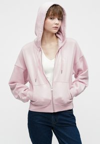 Felpa con cappuccio rosa chiaro con cerniera, cappuccio ampio, maniche lunghe e tasche frontali. Realizzata in materiale morbido con una texture liscia.