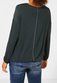 Dark green long-sleeve top with a rounded neckline, featuring a central seam detail at the back and relaxed fit. Paired with blue jeans.