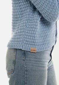 Lichtblauwe gebreide trui met een gestructureerd patroon, voorzien van een geribde zoom en een klein leren label op de onderkant links. Gecombineerd met blauwe jeans.