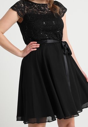 Mujer con un vestido negro con cuerpo de encaje y lentejuelas, mangas cortas transparentes y un lazo de satén atado en la cintura.