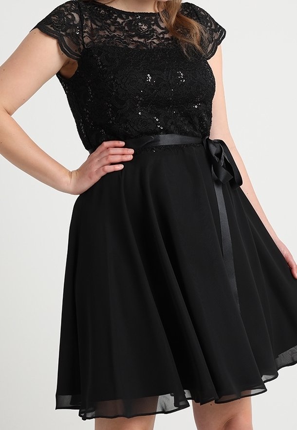 Femme portant une robe noire avec un corsage en dentelle et paillettes, manches courtes transparentes, et un ruban en satin noué à la taille.
