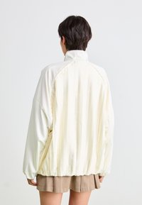 Giubbotto imbottito in crema chiaro con un design della schiena plissettato. Presenta un colletto alto e una vita elasticizzata, realizzato in tessuto liscio con una finitura opaca.