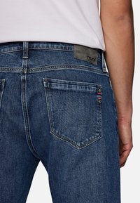 Lähikuva henkilöstä, joka käyttää sinisiä farkkuja, joissa on näkyvä takatasku ja musta "mavi"-merkki vyötäröllä.