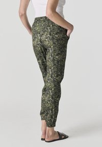 Femme portant un pantalon de jogging vert à motif floral avec des poignets élastiques et des sandales slide noires, debout les mains dans les poches, vue de dos.