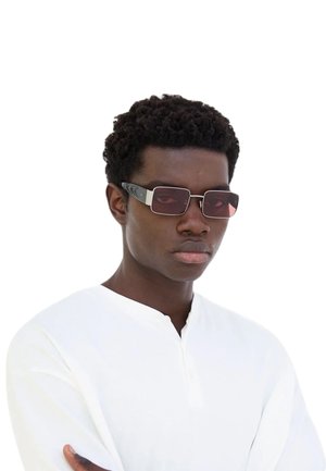 Gli occhiali da sole presentano una montatura rettangolare, sottili aste scure e lenti rosse oscurate. Il modello indossa una maglietta bianca a maniche lunghe, con le braccia incrociate.