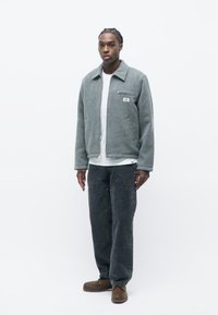 Gray wool jacket with a zip front and pockets, paired with a white shirt and black denim pants. Model stands against a plain white background.