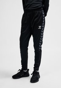 Osoba ubrana w czarne sportowe spodnie z wzorzystymi paskami po bokach oraz czarne buty sportowe, stojąca na jednolitym jasnym tle.