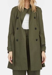 Olijfgroene trenchcoat met een dubbelzijdig ontwerp, zwarte knopen en een iets oversized pasvorm. De stof lijkt glad en licht van gewicht.