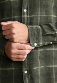 Camisa de cuadros verdes de botones, hecha de un tejido suave, cuenta con botones blancos, un cuello y detalles en los puños con costuras en contraste.
