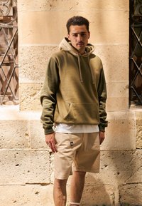 Olive green and beige hoodie with front pocket and drawstrings, paired with beige shorts featuring a logo patch and light texture.