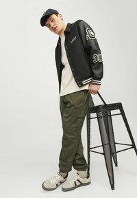 Chaqueta de béisbol negra con mangas de cuero, rayas blancas, camiseta blanca, pantalones cargo color oliva, zapatillas grises, sosteniendo un taburete de metal negro.