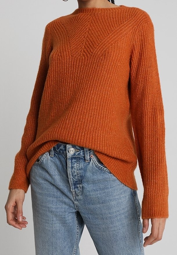 Pull en tricot orange avec un col rond, texture côtelée et fentes sur les côtés, associé à un jean bleu clair avec une fermeture à bouton.