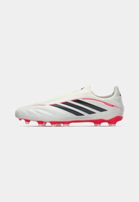 Λευκά ποδοσφαιρικά παπούτσια με μαύρες ρίγες και κόκκινες λεπτομέρειες, με αεροδυναμικό σχέδιο και υφή επιφάνειας για πρόσφυση και απόδοση.