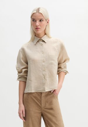Blonde woman wearing loose beige button-up shirt with rolled-up sleeves and brown pants, standing against plain white background.