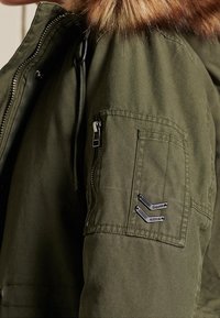 Manica di giacca verde oliva con tasca con zip e due spille a forma di chevron in metallo, caratterizzata da un bordo in pelliccia sintetica marrone.