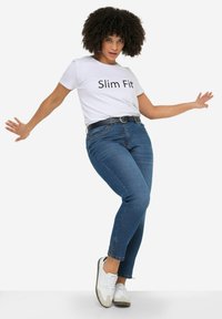 Camiseta blanca ajustada con texto negro, jeans azul oscuro y zapatillas blancas. El modelo posa con los brazos extendidos frente a un fondo blanco.
