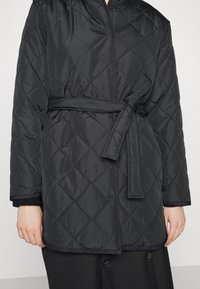 Veste matelassée noire avec ceinture nouée à la taille, manches longues, et texture légèrement rembourrée sur le torse d'une personne.