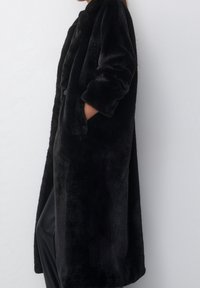 Lungo cappotto nero in pelliccia sintetica con trama morbida, caratterizzato da una vestibilità rilassata, colletto con revers e due tasche laterali. Dettaglio con chiusura a bottoni visibile.
