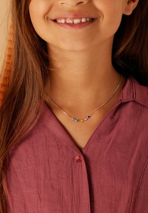 Zilveren ketting met kleurrijke edelsteenaccenten in een gebogen vorm, gedragen door een kind met lang bruin haar, gecombineerd met een gestructureerd bordeauxrood shirt.