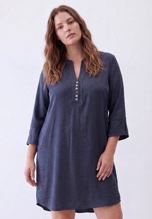 Woman with wavy hair wearing a loose-fitting dark blue dress with three-quarter sleeves and six buttons on the front placket.