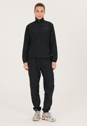 Zwarte lichtgewicht jas met een hoge kraag, ritsluiting en elastische manchetten, gecombineerd met bijpassende zwarte broek en grijze sportschoenen.