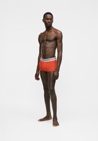 Schlanker dunkelhäutiger Mann, barfuß stehend, trägt rote Boxershorts mit weiß-grauem elastischem Bund vor einfachem weißem Hintergrund.