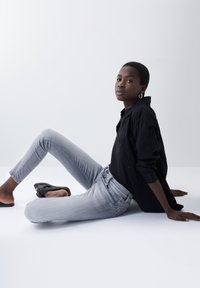 Schwarzes Button-Up-Hemd, graue Skinny Jeans und schwarze Slipper. Modell sitzt mit ausgestreckten Beinen und Händen auf dem Boden.