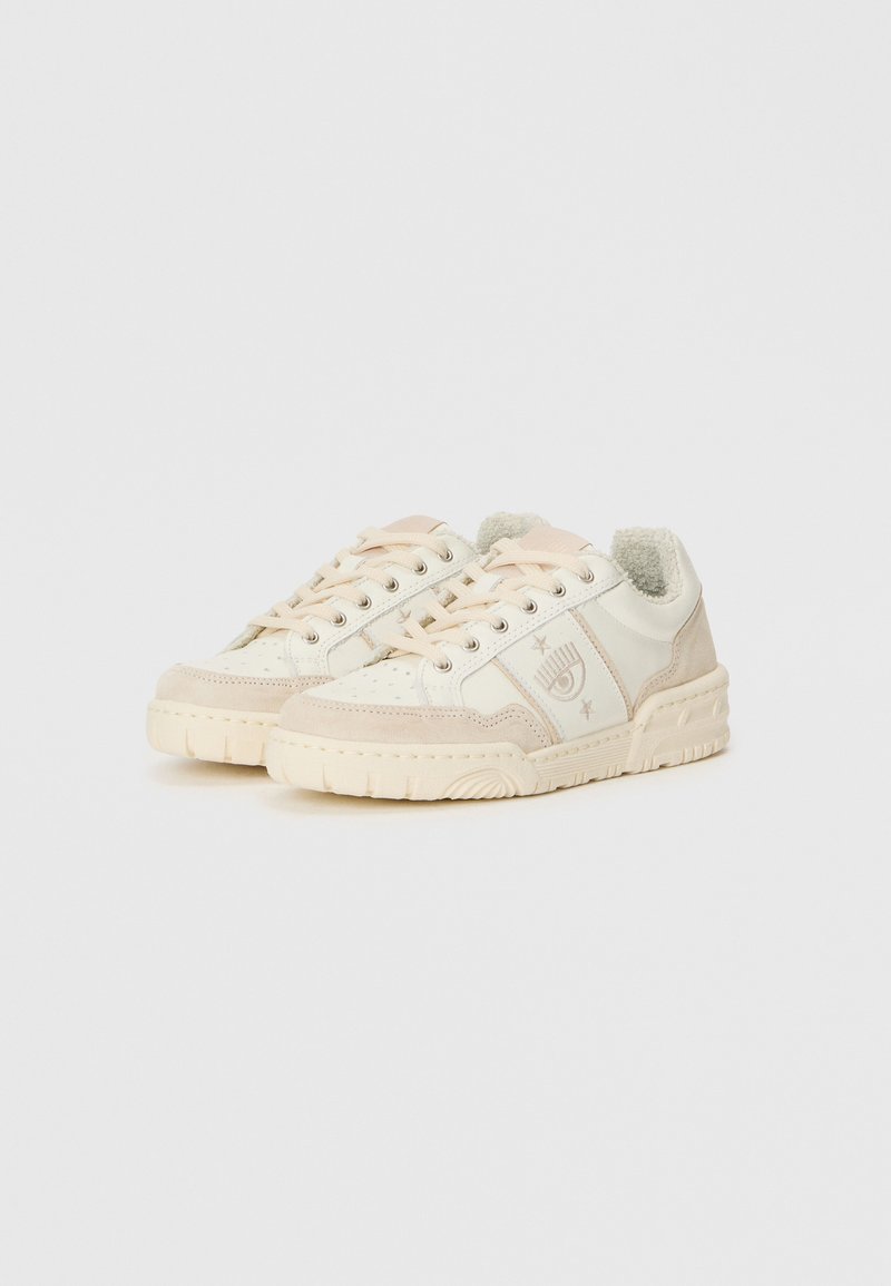Zapatillas bajas crema con detalles de ante beige, cordones y logo de ojo con estrellas de lado, presentadas sobre fondo blanco.