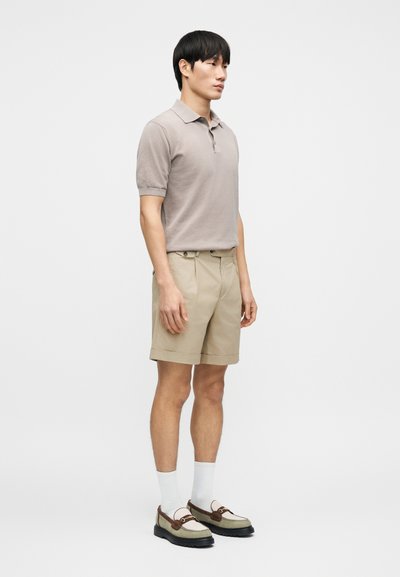 Μπεζ πόλο πουκάμισο, μπεζ σορτς μέχρι το γόνατο, λευκές κάλτσες και παπούτσια με καφέ λεπτομέρειες. Το μοντέλο στέκεται στο πλάι μπροστά σε λευκό φόντο.