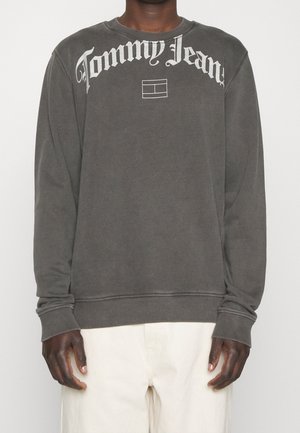 Person trägt einen dunkelgrauen Sweatshirt mit langen Ärmeln mit dem "Tommy Jeans" Logo und einem rechteckigen Emblem auf der Brust, kombiniert mit hellen Hosen.