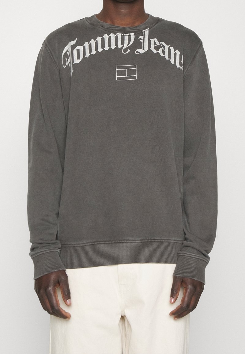 Personne portant un sweat-shirt à manches longues gris foncé avec le logo « Tommy Jeans » et un emblème rectangulaire sur la poitrine, associé à un pantalon de couleur claire.