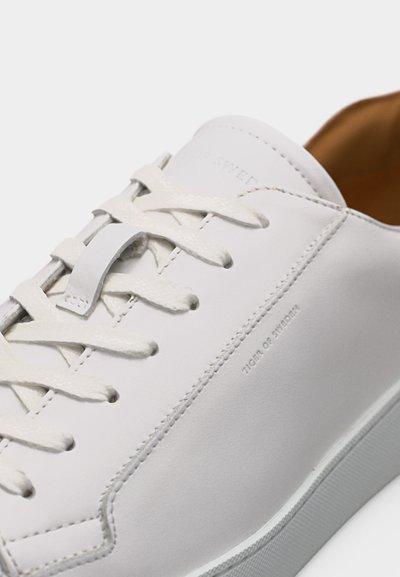 Balto odos sportbačiai su baltais raišteliais, matomi siūlės ir išraižytas užrašas „Tiger of Sweden“ ant šono, rodoma iš arti.