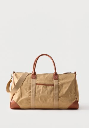 Béžová plátená športová taška s hnedými koženými rúčkami a ozdobami na rohoch, vybavená odnímateľným ramenným popruhom a predným vreckom so zipsom.