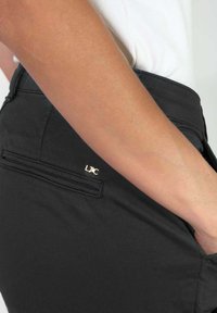 Zwarte broek met een gouden logo-accent, slim fit en een gladde textuur, voorzien van een achterzakdetail.