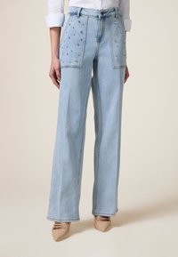 Pantalones vaqueros de pierna ancha de color azul claro con bolsillos adornados con tachuelas metálicas. Diseño clásico de cuatro bolsillos y trabillas para cinturón en la cintura.