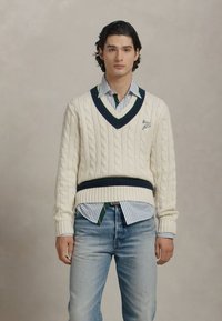Crème kabelgebreide trui met een marineblauwe v-hals en tailleband, gedragen over een lichtblauw en wit gestreept shirt en gecombineerd met lichtblauwe spijkerbroek.