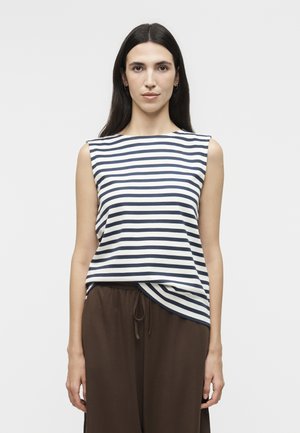 Γυναίκα με μακριά σκούρα μαλλιά που φοράει αμάνικο μπλε σκούρο και λευκό ριγέ τοπ και φαρδιά καφέ παντελόνια, στέκεται μπροστά σε απλό φόντο.