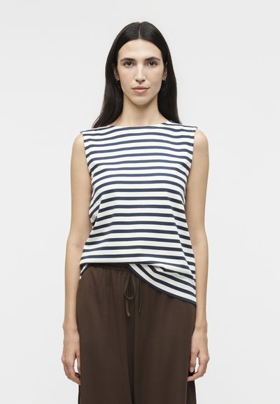 Woman with long dark hair wearing a sleeveless navy and white striped top and loose brown pants, standing against a plain background.